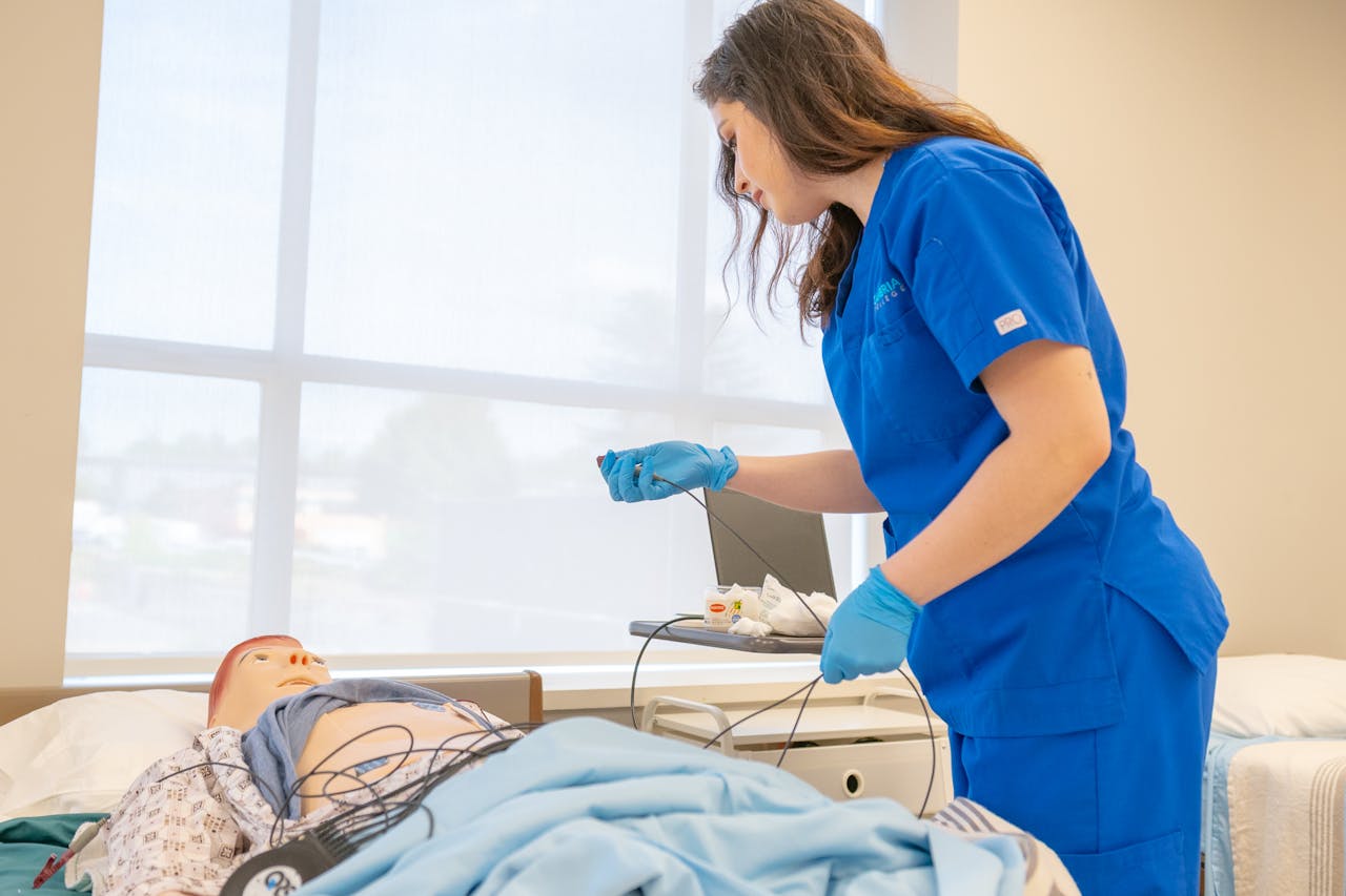 A nursing student in blue scrubs attends to a medical mannequin in a training room.